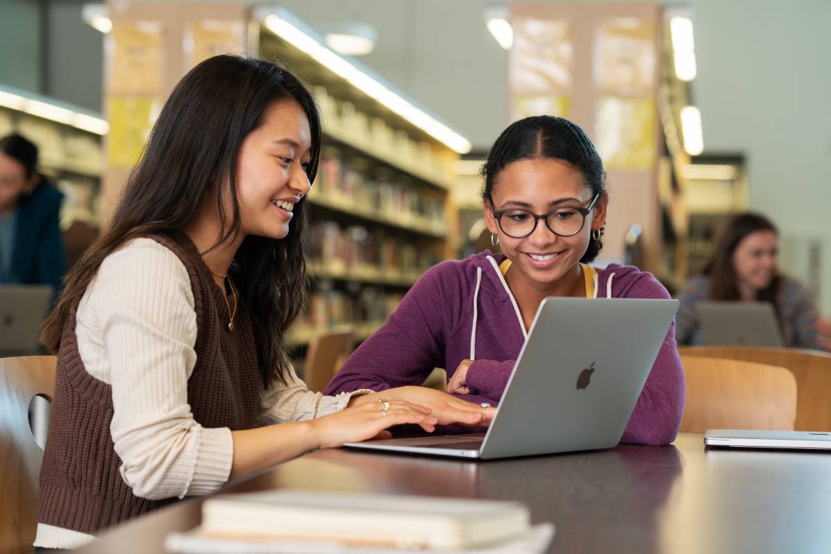 Zwei Schülerinnen sitzen mit einem MacBook in einer Bibliothek